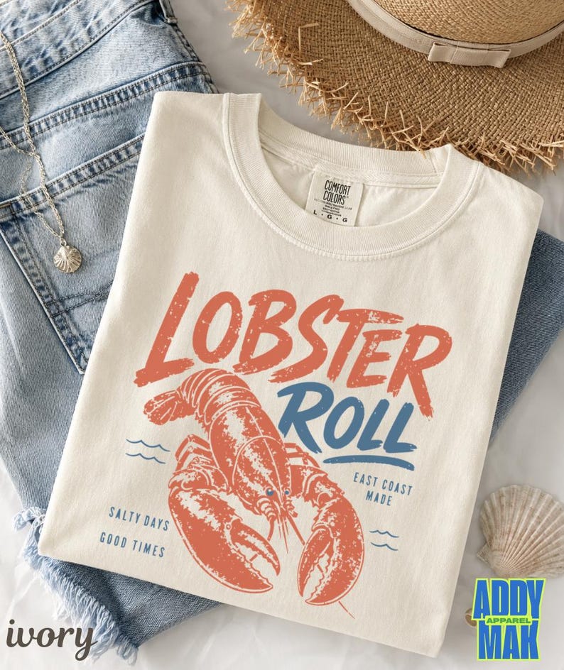 Pu&ograve; includere: T-shirt color avorio con grafica arancione invecchiata con la scritta "Lobster Roll" sopra un'illustrazione dettagliata di un'aragosta. La maglietta presenta anche il testo "East Coast Made" e "Salty Days Good Times".