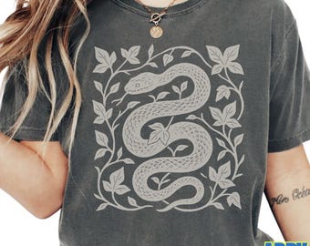 May include: Dark gray t-shirt featuring a detailed, light gray illustration of a snake coiled within a square frame of leaves and vines. The word "pepper" is printed at the bottom left.