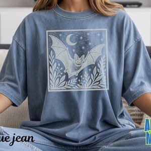 May include: A blue denim-colored t-shirt featuring a square graphic print on the chest. The design depicts a bat in flight surrounded by foliage, a crescent moon, and stars. The shirt has a crew neckline, short sleeves, and a relaxed fit.