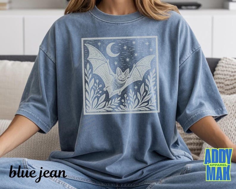 May include: A blue denim-colored t-shirt featuring a square graphic print on the chest. The design depicts a bat in flight surrounded by foliage, a crescent moon, and stars. The shirt has a crew neckline, short sleeves, and a relaxed fit.