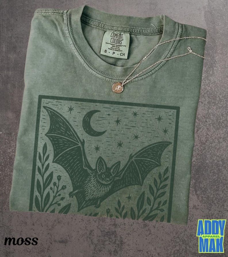 May include: A sage green short-sleeve t-shirt featuring a dark green graphic print of a bat in flight under a crescent moon and stars, framed by foliage. A delicate silver-tone chain necklace with a small round pendant rests on the neckline.