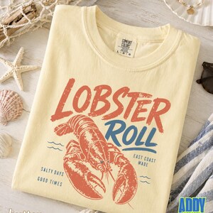 Pu&ograve; includere: T-shirt color burro con grafica rossa e blu invecchiata con la scritta "Lobster Roll". Il design presenta un'illustrazione di aragosta e le frasi "Salty Days, Good Times" e "East Coast Made."