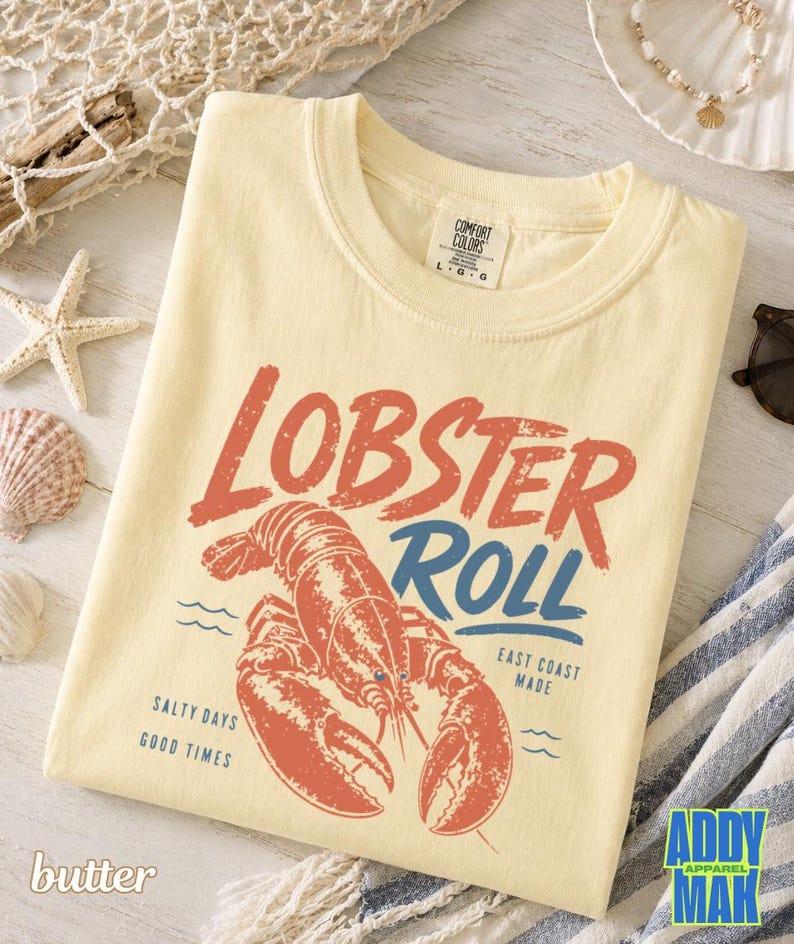 Pu&ograve; includere: T-shirt color burro con grafica rossa e blu invecchiata con la scritta "Lobster Roll". Il design presenta un'illustrazione di aragosta e le frasi "Salty Days, Good Times" e "East Coast Made."