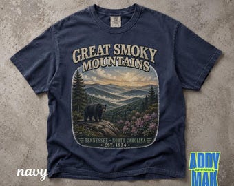 May include: A navy blue short-sleeve t-shirt featuring a graphic print of the Great Smoky Mountains. The design includes a black bear on a rocky outcrop, pine trees, flowering bushes, and the text Great Smoky Mountains Tennessee North Carolina Est. 1934.