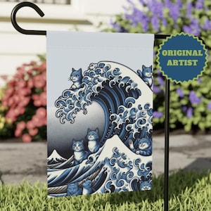 May include: A garden flag featuring a blue and white illustration of cats riding a large ocean wave, with the text "ORIGINAL ARTIST" on a blue badge. The flag is displayed outdoors on a black metal stand.