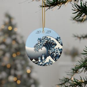 May include: Round Christmas ornament with a blue and white design featuring cats in a wave, reminiscent of Hokusai's "The Great Wave off Kanagawa." The ornament has the year "2025" printed on it. It is hanging from a gold string.