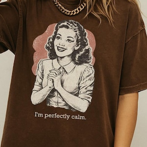 Op de afbeelding: Bruin t-shirt met een retro illustratie van een vrouw en de zin "I'm perfectly calm." De afbeelding staat tegen een roze achtergrond. Het shirt heeft korte mouwen en een losse pasvorm. De merknaam "Addy Mak" is zichtbaar.