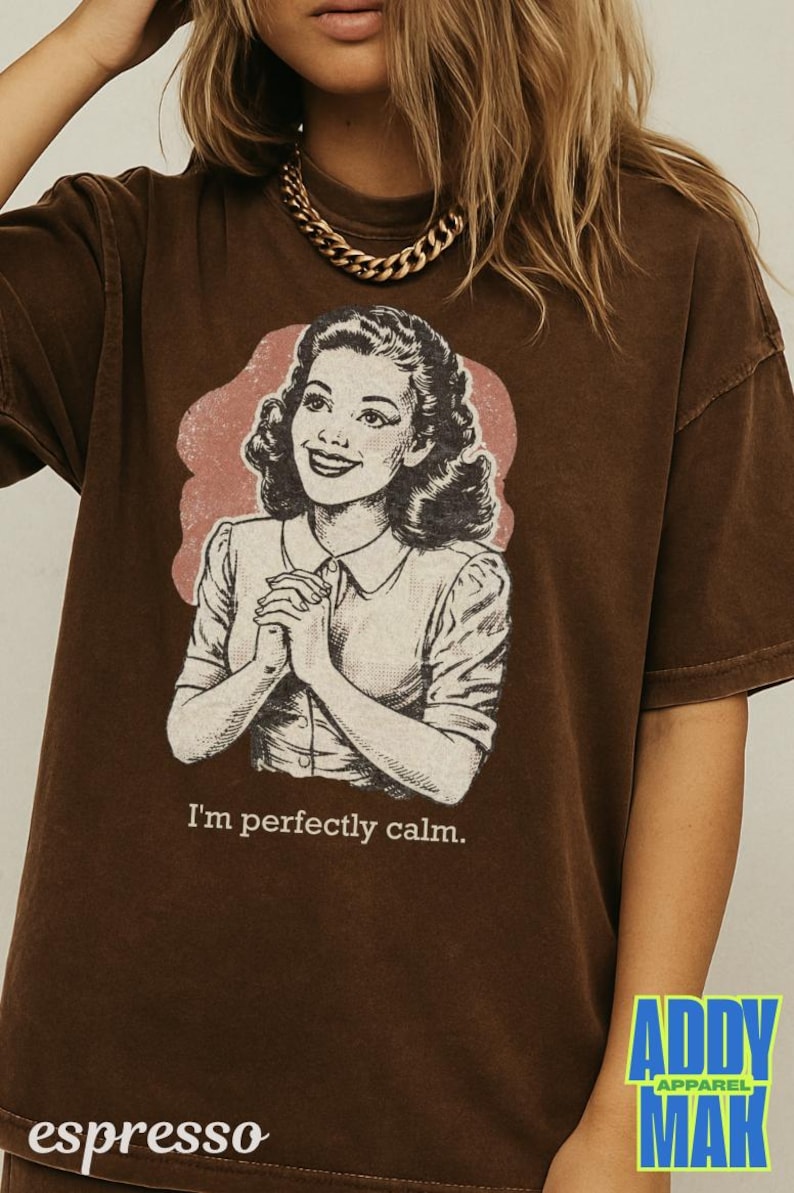 Op de afbeelding: Bruin t-shirt met een retro illustratie van een vrouw en de zin "I'm perfectly calm." De afbeelding staat tegen een roze achtergrond. Het shirt heeft korte mouwen en een losse pasvorm. De merknaam "Addy Mak" is zichtbaar.