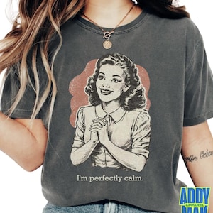 Op de afbeelding: Donkergrijs t-shirt met een vintage illustratie van een vrouw en de tekst "I'm perfectly calm." De afbeelding is in retrostijl met een rode achtergrond. Het shirt heeft korte mouwen en een ronde hals.