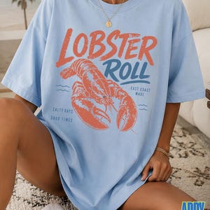 Pu&ograve; includere: T-shirt azzurra con una grafica di un'aragosta e le parole "Lobster Roll" in arancione e blu. La maglietta presenta anche le frasi "East Coast Made" e "Salty Days Good Times".