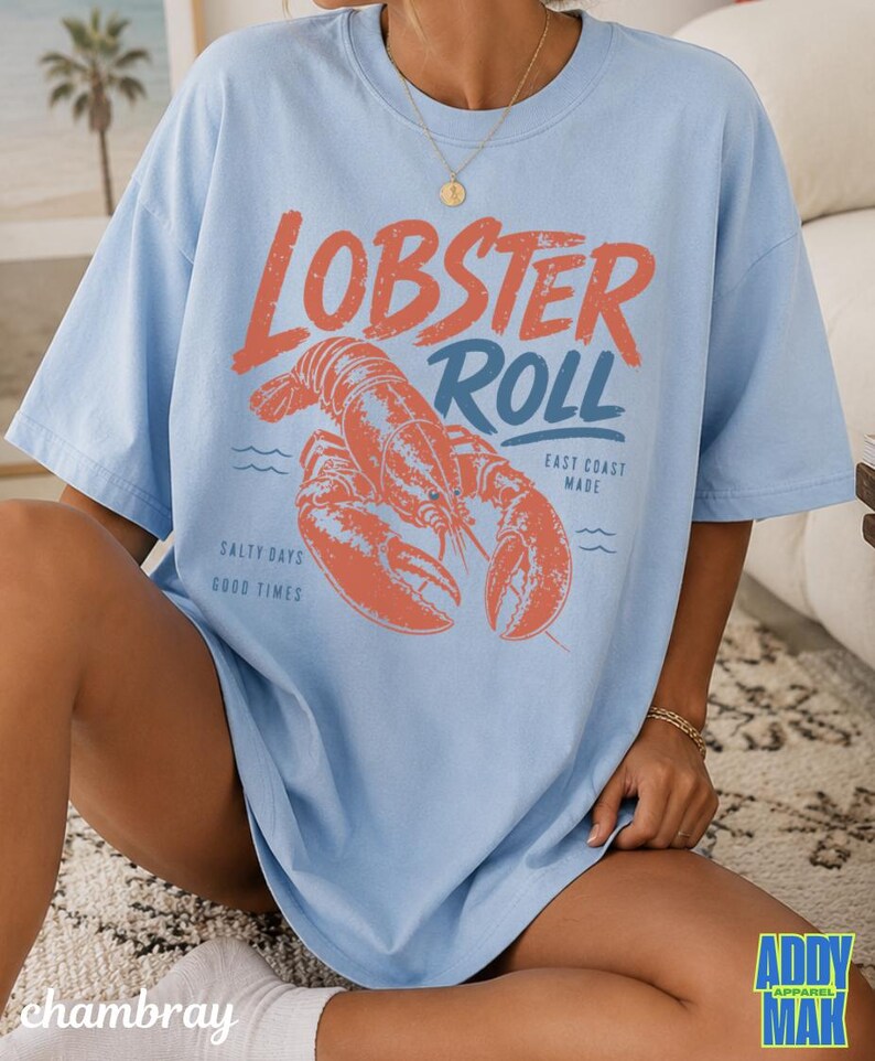 Pu&ograve; includere: T-shirt azzurra con una grafica di un'aragosta e le parole "Lobster Roll" in arancione e blu. La maglietta presenta anche le frasi "East Coast Made" e "Salty Days Good Times".