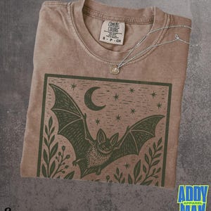 May include: A brown short-sleeve t-shirt featuring a dark green graphic print of a bat in flight under a crescent moon and stars, framed by foliage. The shirt has a crew neckline and a Comfort Colors brand label inside the collar.