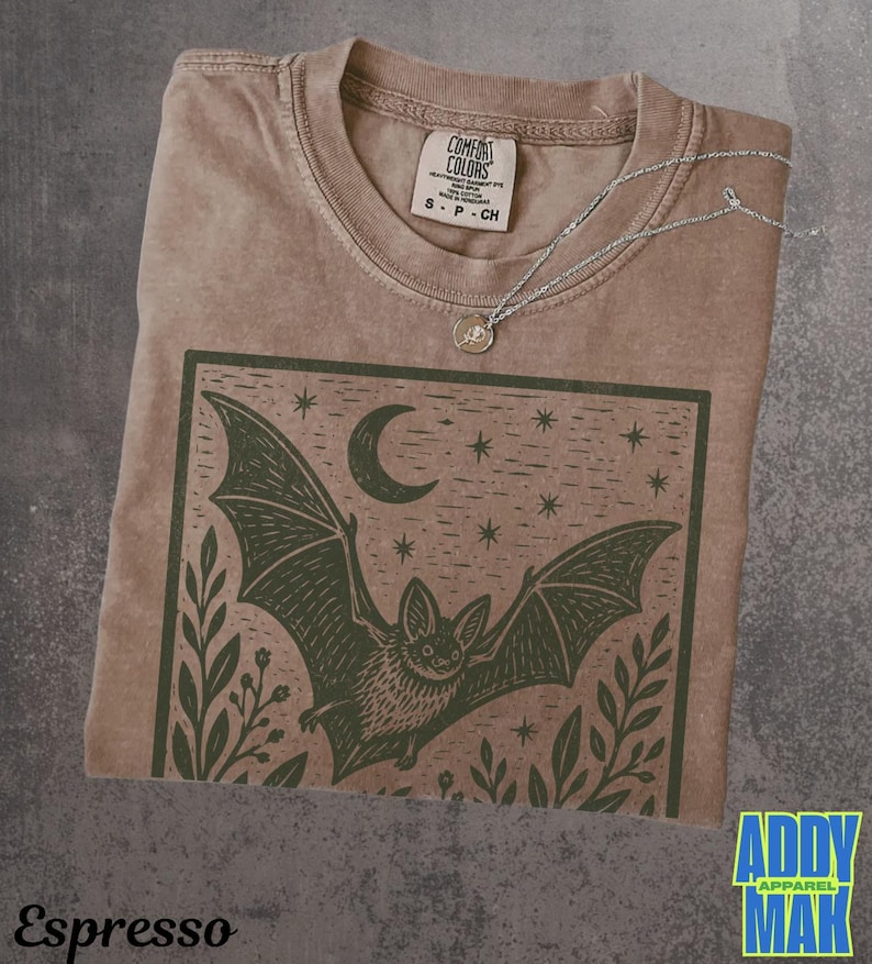 May include: A brown short-sleeve t-shirt featuring a dark green graphic print of a bat in flight under a crescent moon and stars, framed by foliage. The shirt has a crew neckline and a Comfort Colors brand label inside the collar.