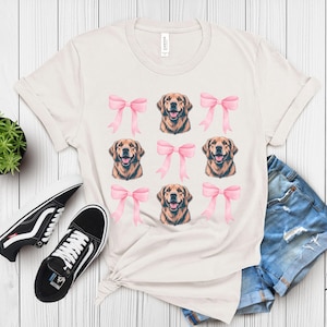 Puede incluir: Camiseta de color crema con un estampado de caras de golden retriever y lazos rosas. La camiseta se combina con zapatillas negras, pantalones cortos vaqueros y una peque&ntilde;a planta en maceta. Una prenda informal con tem&aacute;tica de perros.