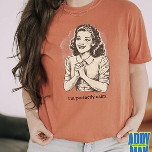 Op de afbeelding: Verbrand oranje t-shirt met een vintage illustratie van een vrouw en de tekst "I'm perfectly calm." De afbeelding is in zwart en wit. Het shirt heeft korte mouwen en een ronde hals.