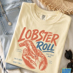 Pu&ograve; includere: T-shirt color crema con grafica rossa e blu con la scritta "Lobster Roll" e un'illustrazione di aragosta. La maglietta riporta anche le frasi "Salty Days Good Times" e "East Coast Made".
