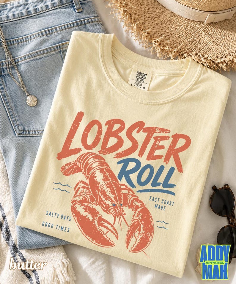 Pu&ograve; includere: T-shirt color crema con grafica rossa e blu con la scritta "Lobster Roll" e un'illustrazione di aragosta. La maglietta riporta anche le frasi "Salty Days Good Times" e "East Coast Made".