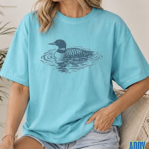 Puede incluir: Una camiseta de manga corta de color turquesa brillante con una ilustraci&oacute;n gr&aacute;fica de color azul marino oscuro de un colimbo nadando en el agua. El dise&ntilde;o est&aacute; centrado en el pecho de la camiseta, que tiene cuello redondo y corte holgado.