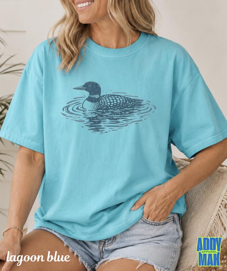 Puede incluir: Una camiseta de manga corta de color turquesa brillante con una ilustraci&oacute;n gr&aacute;fica de color azul marino oscuro de un colimbo nadando en el agua. El dise&ntilde;o est&aacute; centrado en el pecho de la camiseta, que tiene cuello redondo y corte holgado.