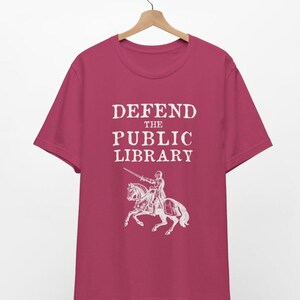 May include: A pink t-shirt with the text "DEFEND THE PUBLIC LIBRARY" in white. A white illustration of a knight on horseback with a sword is below the text.