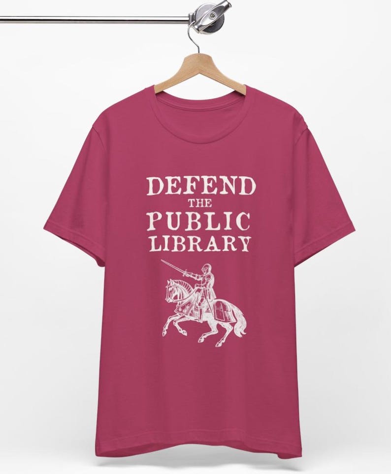 May include: A pink t-shirt with the text "DEFEND THE PUBLIC LIBRARY" in white. A white illustration of a knight on horseback with a sword is below the text.