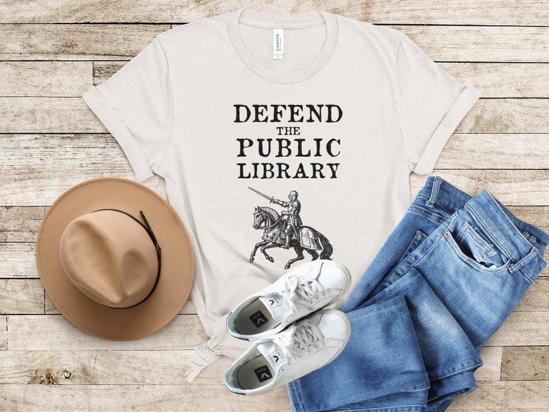 May include: A white t-shirt with the text "DEFEND THE PUBLIC LIBRARY" and a black and white illustration of a knight riding a horse. The shirt is laid out on a wooden surface with a brown hat and blue jeans.
