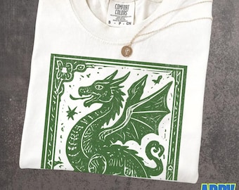 May include: An ivory t-shirt featuring a green block-print style graphic of a dragon within a rectangular border. The shirt has a crew neckline and a Comfort Colors brand label at the inner collar. A thin gold chain necklace rests on the fabric.