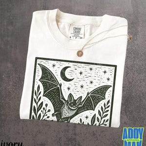 May include: An ivory short-sleeve t-shirt featuring a dark green block print graphic of a bat in flight under a crescent moon and stars, framed by foliage. The inner neck label reads Comfort Colors, size S, 100% cotton, made in Honduras.