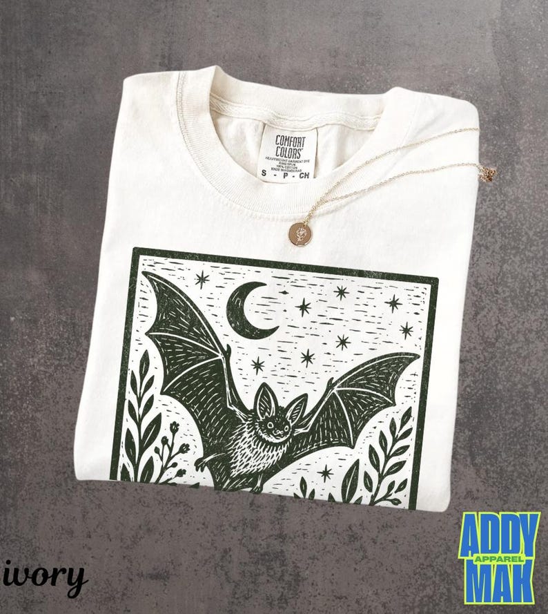 May include: An ivory short-sleeve t-shirt featuring a dark green block print graphic of a bat in flight under a crescent moon and stars, framed by foliage. The inner neck label reads Comfort Colors, size S, 100% cotton, made in Honduras.