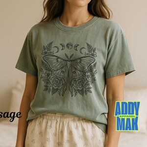 May include: Sage green t-shirt featuring a detailed black illustration of a moth with floral and moon phase elements. The shirt has short sleeves and a crew neck. The word "sage" is printed on the left side of the shirt.