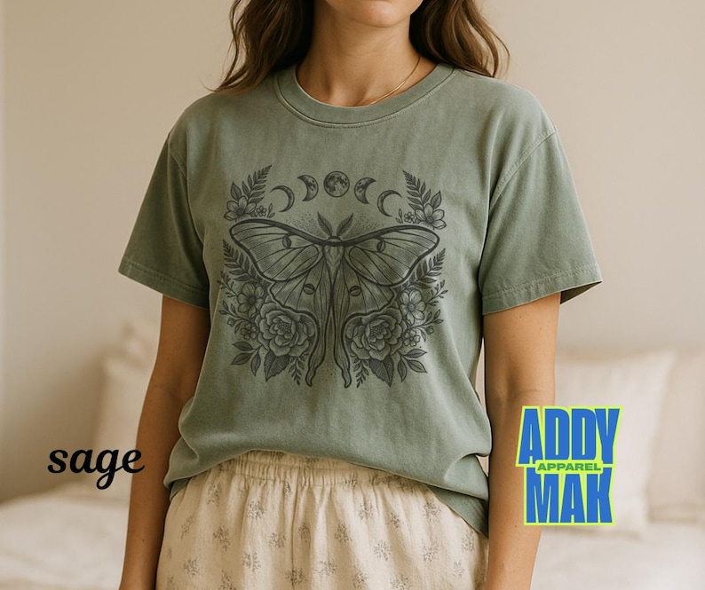 May include: Sage green t-shirt featuring a detailed black illustration of a moth with floral and moon phase elements. The shirt has short sleeves and a crew neck. The word "sage" is printed on the left side of the shirt.
