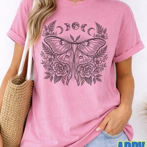 May include: Pink t-shirt with a large black and white moth graphic, surrounded by floral and moon phase designs. The shirt has rolled-up sleeves and the word "blossom" is printed on the bottom left. The brand name "ADDY MAK" is printed on the bottom right.