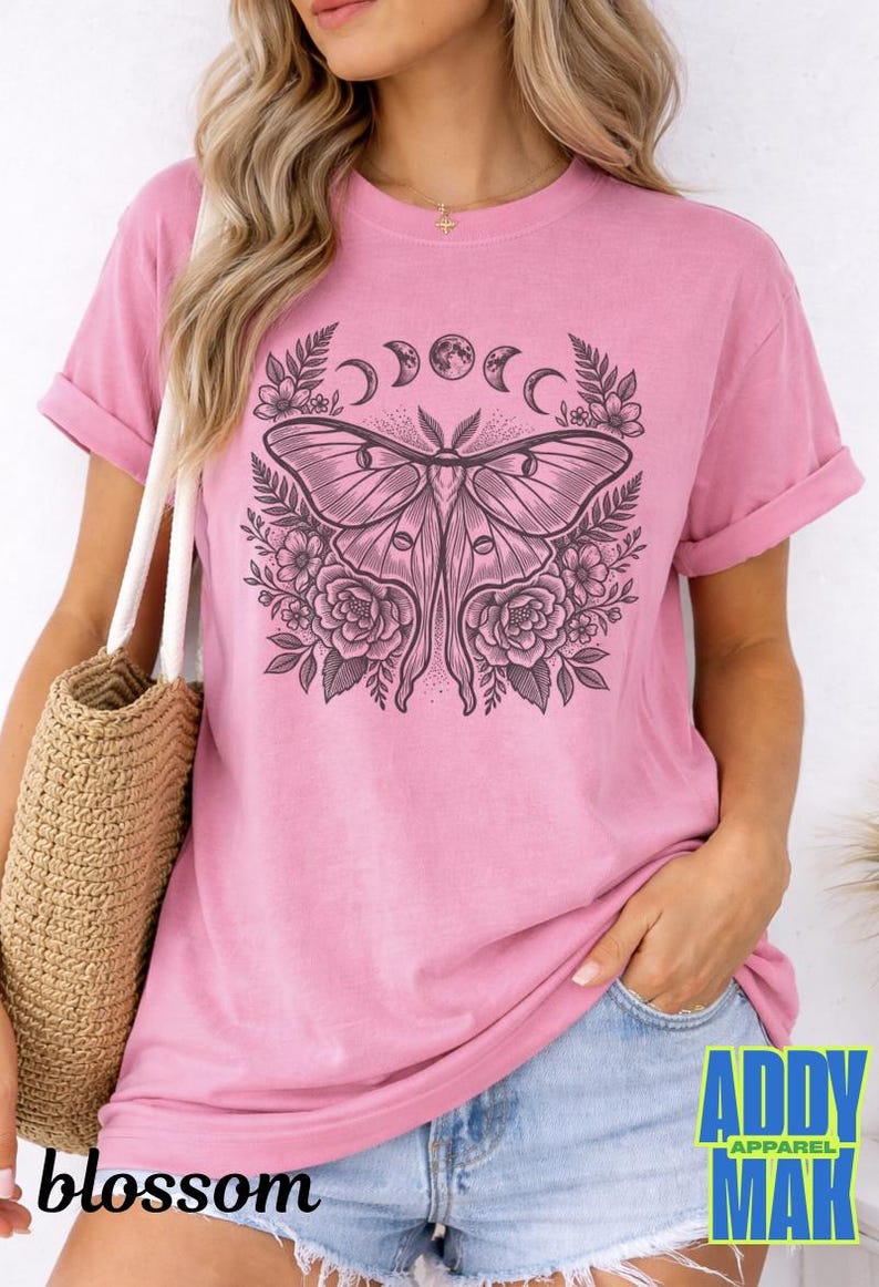 May include: Pink t-shirt with a large black and white moth graphic, surrounded by floral and moon phase designs. The shirt has rolled-up sleeves and the word "blossom" is printed on the bottom left. The brand name "ADDY MAK" is printed on the bottom right.