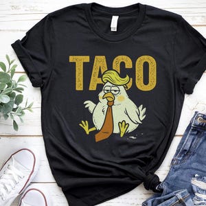 May include: Black t-shirt with a humorous graphic of a chicken wearing a tie and a blonde comb-over, with the word "TACO" above it. The shirt is paired with blue jeans and white sneakers.