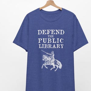 May include: A royal blue t-shirt with the text "DEFEND THE PUBLIC LIBRARY" in white, featuring a knight on horseback with a sword.