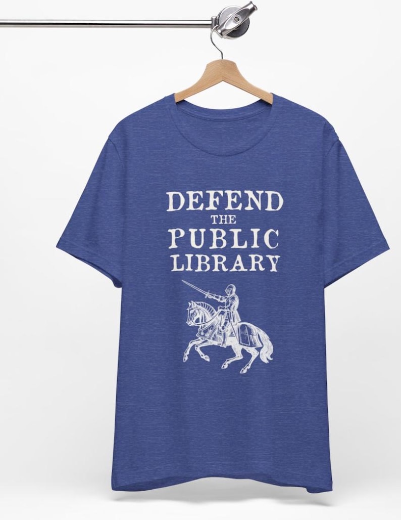 May include: A royal blue t-shirt with the text "DEFEND THE PUBLIC LIBRARY" in white, featuring a knight on horseback with a sword.