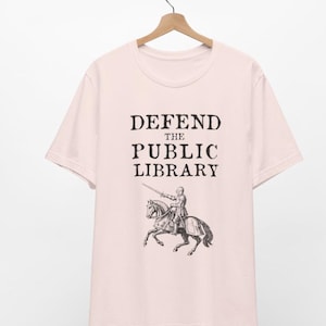 May include: A pink t-shirt with the text "DEFEND THE PUBLIC LIBRARY" and a black and white illustration of a knight on horseback.