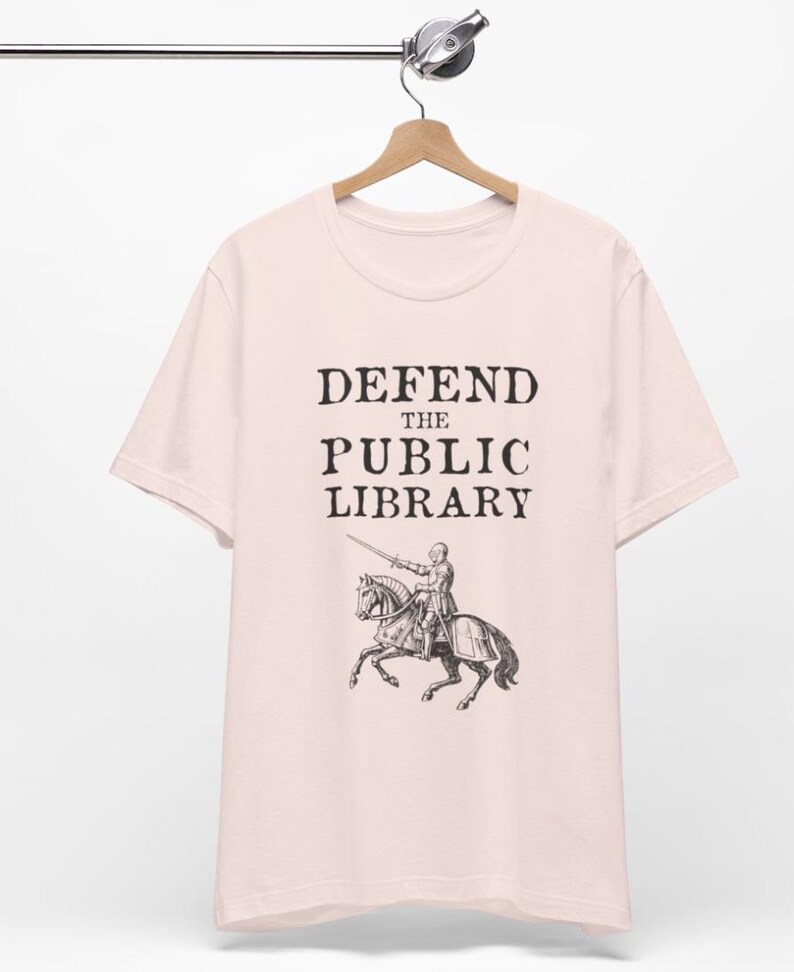 May include: A pink t-shirt with the text "DEFEND THE PUBLIC LIBRARY" and a black and white illustration of a knight on horseback.