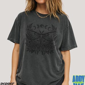 May include: Dark grey t-shirt featuring a detailed black moth design with floral and crescent moon accents. The shirt has a vintage, washed look and the word "pepper" is printed on the bottom left.
