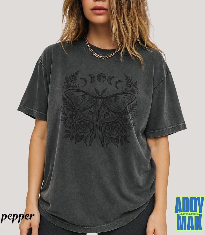 May include: Dark grey t-shirt featuring a detailed black moth design with floral and crescent moon accents. The shirt has a vintage, washed look and the word "pepper" is printed on the bottom left.