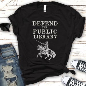 May include: Black t-shirt with white text that reads "Defend the Public Library" and a graphic of a knight on horseback.