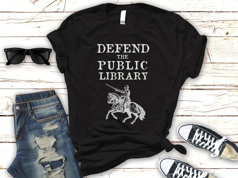 May include: Black t-shirt with white text that reads "Defend the Public Library" and a graphic of a knight on horseback.