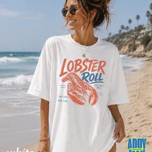 Pu&ograve; includere: T-shirt bianco con grafica rossa e blu "Lobster Roll" e illustrazione di un'aragosta. La maglietta presenta anche il testo "East Coast Made", "Salty Days" e "Good Times".