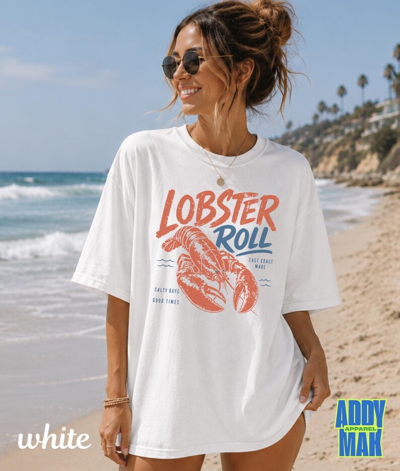 Pu&ograve; includere: T-shirt bianco con grafica rossa e blu "Lobster Roll" e illustrazione di un'aragosta. La maglietta presenta anche il testo "East Coast Made", "Salty Days" e "Good Times".