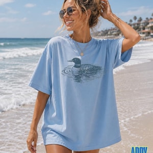Puede incluir: Una camiseta de manga corta de color azul claro con un estampado gr&aacute;fico en azul oscuro de un colimbo nadando en el agua. La prenda tiene cuello redondo y un corte holgado. El texto "chambray" aparece en blanco en la esquina inferior izquierda.