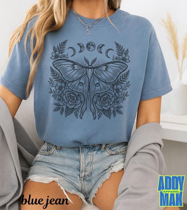 May include: A blue jean colored t-shirt featuring a detailed illustration of a moth with floral and moon phase elements. The graphic is centered on the shirt. The text "blue jean" is at the bottom left of the image, and the brand logo is at the bottom right.