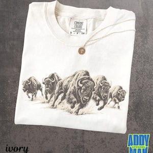 May include: An ivory-colored short-sleeve t-shirt featuring a detailed brown graphic print of a herd of five bison running across the chest. The shirt has a Comfort Colors brand label at the inner neckline and is paired with a thin gold-tone necklace.