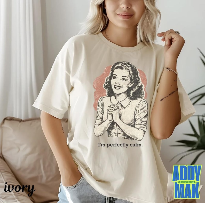 Op de afbeelding: Ivoor kleurig T-shirt met een vintage illustratie van een vrouw en de tekst "I'm perfectly calm." De afbeelding staat tegen een roze achtergrond. Het shirt heeft korte mouwen en een relaxte pasvorm. Het merklogo "Addy Mak Apparel" is zichtbaar.