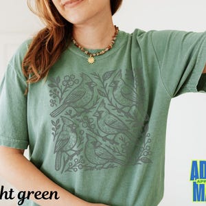 May include: Light green t-shirt featuring a gray cardinal bird design. The graphic includes several birds, branches, and berries. The words "light green" are printed at the bottom left. The brand logo "ADDY MAK" is at the bottom right.