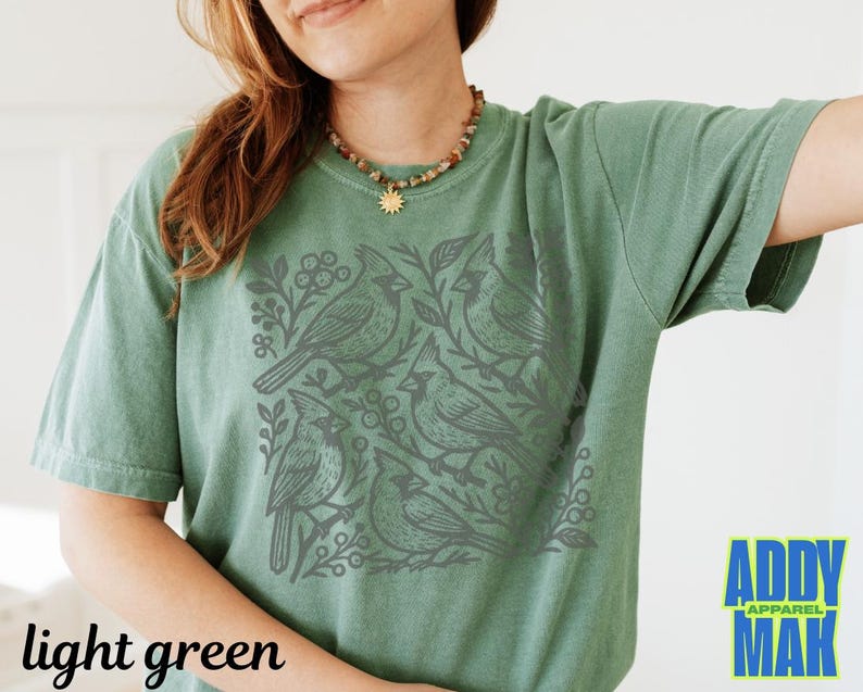 May include: Light green t-shirt featuring a gray cardinal bird design. The graphic includes several birds, branches, and berries. The words "light green" are printed at the bottom left. The brand logo "ADDY MAK" is at the bottom right.